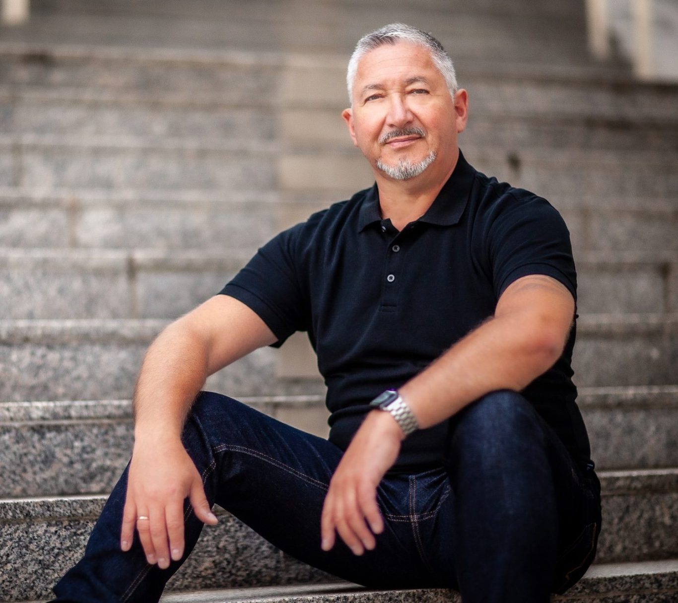 Trauerredner Frank Mohr, bekleidet mit dunkelblauer Jeans und schwarzem Poloshirt, sitzt auf einer Treppe, schaut freundlich in die Kamera.